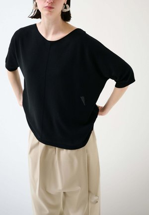 Donna che indossa una maglia nera larga a maniche corte, pantaloni beige a vita alta e larghi, con grandi orecchini quadrati, in piedi con le mani sui fianchi.