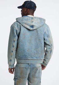 Giacca di jeans azzurro chiaro con cappuccio, caratterizzata da una texture leggermente scolorita e polsini a costine. Indossata con pantaloni di jeans azzurro chiaro abbinati.
