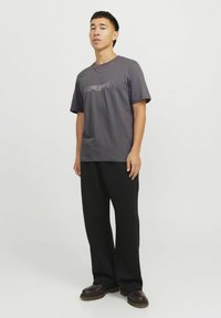 Donkergrijze t-shirt met korte mouwen met grafische tekst op de borst, gecombineerd met zwarte wijde broek en bruine schoenen. Gladde katoenstructuur.