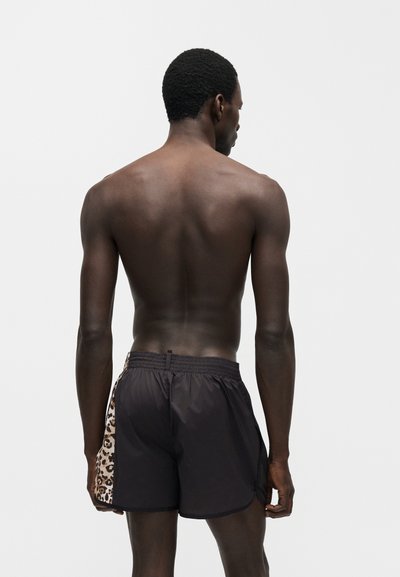Νέος άντρας που φαίνεται από πίσω, χωρίς πουκάμισο, φοράει μαύρο μαγιό με πάνελ λεοπάρδαλης στη μία πλευρά.