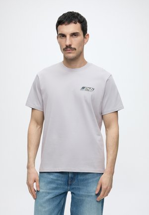 Mann mit kurzen dunklen Haaren und Schnurrbart, der ein hellgraues T-Shirt mit kleinem Logo auf der Brust und blaue Jeans trägt, steht vor einem einfarbigen Hintergrund.