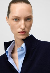 Maglione lavorato a maglia navy con colletto alto indossato sopra una camicia azzurro chiaro con bottoni. Texture liscia, vestibilità su misura e colori a contrasto.