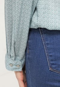 Blouse vert clair avec un petit motif floral blanc, poignets boutonnés et tissu doux, assortie à un jean en denim bleu foncé.
