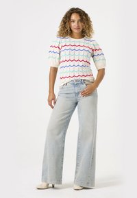 Gebreide top met zigzagpatroon in rood, blauw en turquoise, gecombineerd met hooggetailleerde lichtblauwe wijde jeans en crèmekleurige schoenen.
