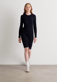 Jūrinės mėlynos tekstūrinės megztos suknelės ilgomis rankovėmis ir apvalia iškirpte, su subtiliu logotipo akcentu krūtinėje. Sukurta su baltais kojiniais ir sportbačiais.