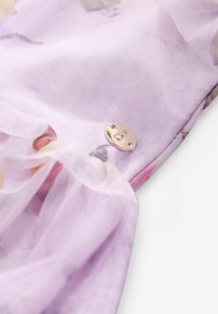 Lila durchsichtiger Stoff mit floralen Mustern, gerafftem Design und einem metallischen Emblem mit dem Buchstaben "B" als Detail.