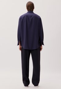 Marineblaues übergroßes Hemd mit langen Ärmeln und seitlichen Schlitzen, aus glattem Stoff. Kombiniert mit locker sitzenden dunklen Hosen.