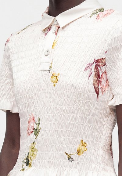 Gros plan d'un top blanc texturé à manches courtes avec tissu smocké, imprimés floraux, col boutonné et bords de manches légèrement froncés.
