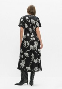 Kobieta ubrana w czarną sukienkę midi z białym kwiatowym wzorem i krótkimi rękawami, zestawiona z czarnymi, spiczastymi kozakami za kolano, stojąca tyłem.