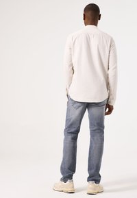 Camisa de manga larga color beige claro con detalles de botones, combinada con jeans azul claro y zapatillas color crema, vista desde la espalda.