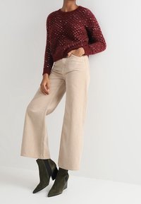 Maglione lavorato a maglia bordeaux con finitura testurizzata, abbinato a pantaloni beige a gamba larga e stivaletti neri con tacco angolare.
