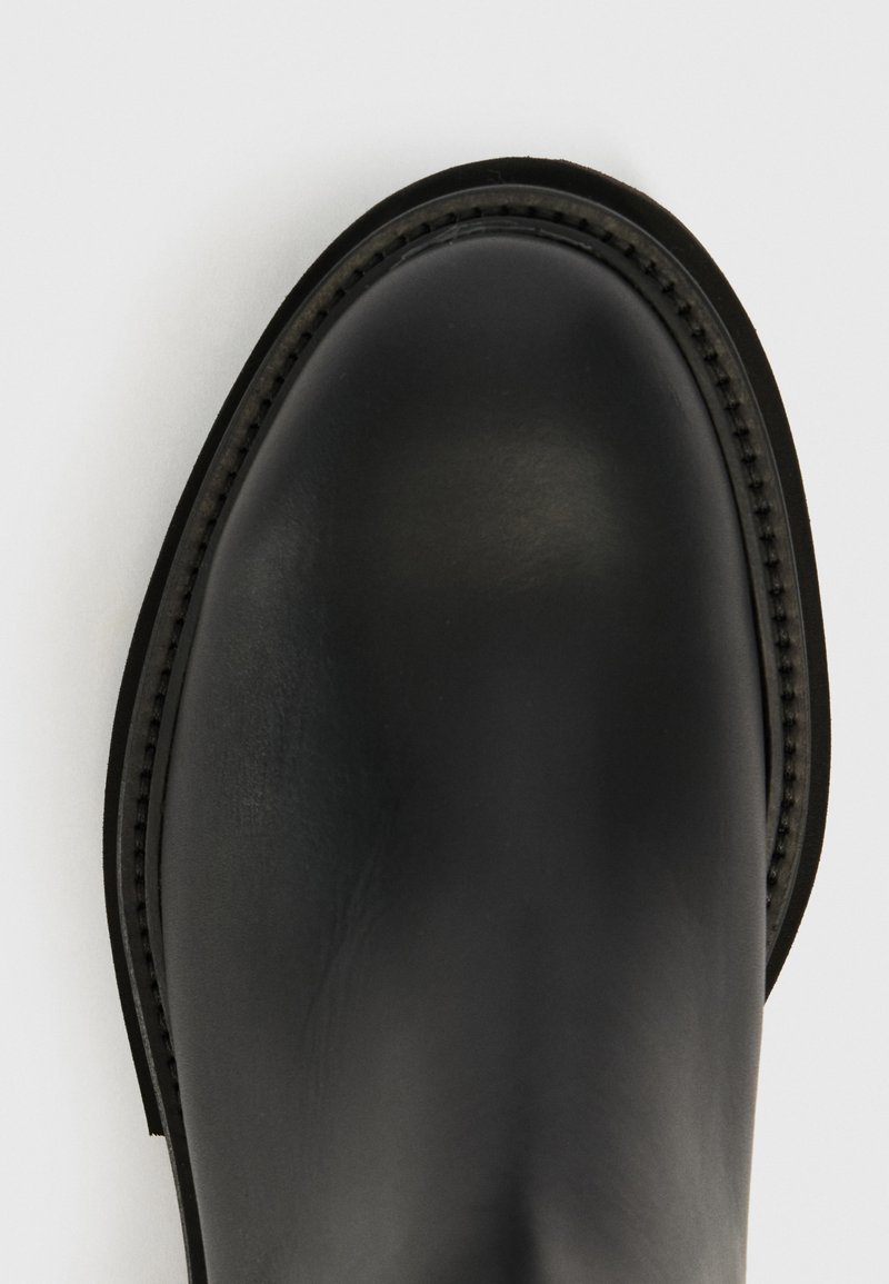 Gros plan sur l'embout arrondi d'une botte en cuir noir avec des coutures détaillées le long du bord de la semelle sur fond blanc.