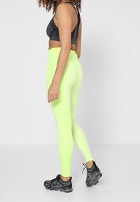 Leggings de mujer de tiro alto en un brillante color amarillo neón, confeccionados con un material elástico. Combinados con un sujetador deportivo de rayas negras y zapatillas negras.
