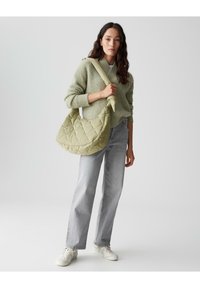 Borsa a spalla trapuntata di colore verde chiaro, dalla forma arrotondata e con una fascia larga. Modello che indossa un maglione a trama, jeans grigi e sneaker bianche.