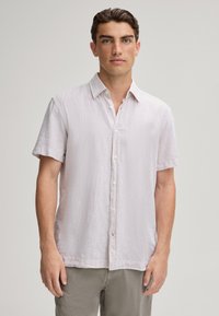 Camisetta grigia chiara a maniche corte in tessuto testurizzato, con bottoni frontali e colletto a punta, abbinata a pantaloni chiari.