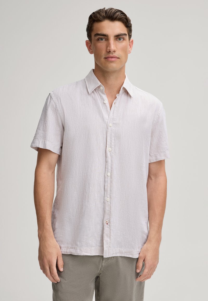 Camisetta grigia chiara a maniche corte in tessuto testurizzato, con bottoni frontali e colletto a punta, abbinata a pantaloni chiari.