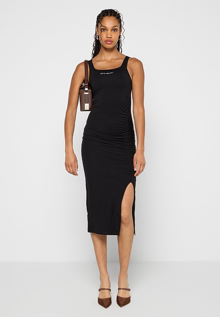 Schwarzes Midi-Kleid mit figurbetontem Design, Trägern, seitlichem Schlitz und Raffung. Getragen mit braunen High Heels und einer kleinen Schultertasche.
