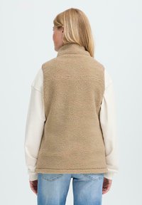 Femme portant un gilet en polaire beige sur un sweat-shirt blanc et un jean bleu, vue de dos contre un fond clair uniforme.