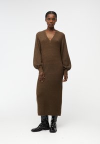 Abito lavorato a maglia marrone con scollatura a V profonda e maniche lunghe a palloncino. Il design presenta una silhouette aderente e una texture a coste. Indossato con stivali neri lucenti.
