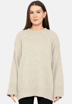 Young woman with straight brown hair wearing a loose beige knit sweater and black pants, standing against a plain white background.