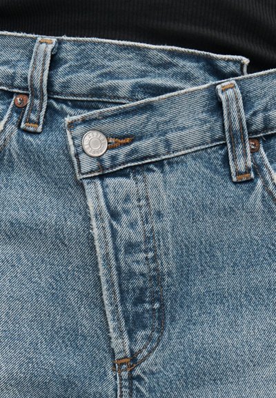 Μπλε τζιν παντελόνι από denim με τη μέση μερικώς ξεκουμπωμένη, που δείχνει τα χαλκόχρωμα καρφιά και τη ραφή σε αντίθεση.