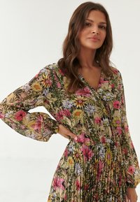 Eine Frau trägt ein langärmeliges Kleid mit Blumenmuster in Pink, Gelb, Weiß und Grün und steht mit einer Hand auf der Hüfte.