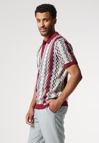 Camisa polo de punto en color crema, con patrones geométricos en rojo y azul. Mangas cortas con cuello y dobladillo de canalé en color marrón. Textura suave.