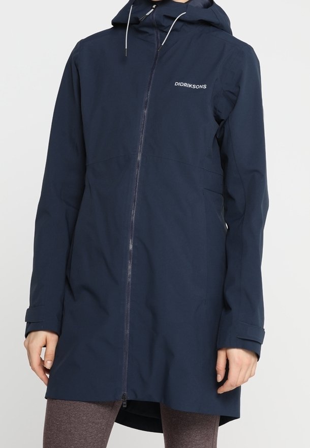 Imperméable bleu marine à capuche avec fermeture éclair frontale, poignets réglables et logo Didriksons sur la poitrine, porté sur des leggings marron.