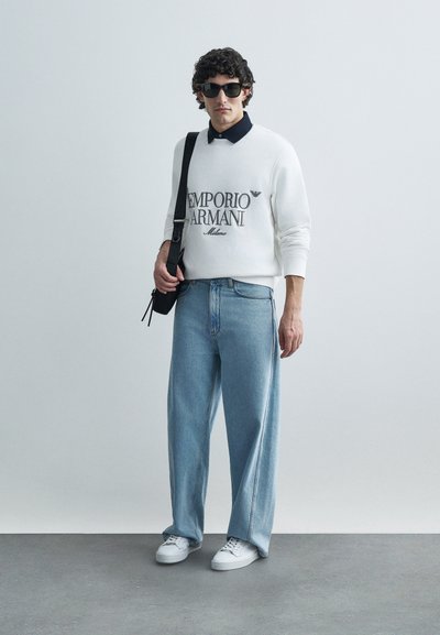 Camisola branca com logótipo preto "Emporio Armani", combinada com jeans largos de lavagem clara e ténis brancos. Acessórios incluem óculos de sol e bolsa pretos.