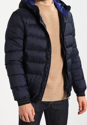 Mand iført en marineblå quiltet jakke med blå hætte, beige ribstrikket sweater og mørke jeans, stående foran en lys baggrund.