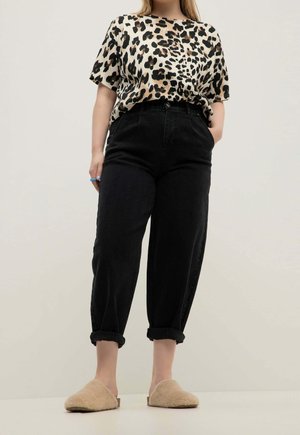 Vrouw draagt een kort mouwtop met luipaardprint, hooggetailleerde zwarte opgerolde broek, beige zachte slippers, staand tegen een lichte achtergrond.