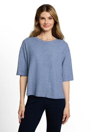 Vrouw met lichtbruin haar die een losse lichtblauwe korte mouwen trui en donkere broek draagt, staand tegen een effen witte achtergrond.