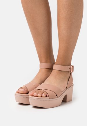 Nude platform sandals with a wide toe strap and ankle buckle, featuring a chunky heel and textured sole for added grip.