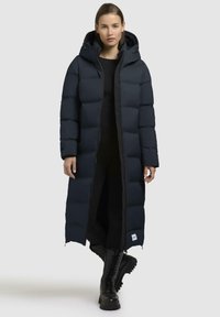 Cappotto lungo imbottito blu navy con cappuccio, dotato di chiusura con zip, design trapuntato e bordi neri. Indossato sopra abbigliamento e stivali neri.