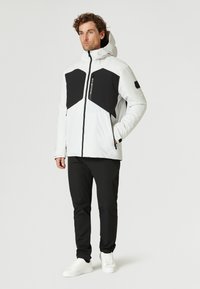 Vit och svart jacka med huva, med ett geometriskt mönster, dragkedja fram och två sidofickor. Parat med svarta byxor och vita sneakers.