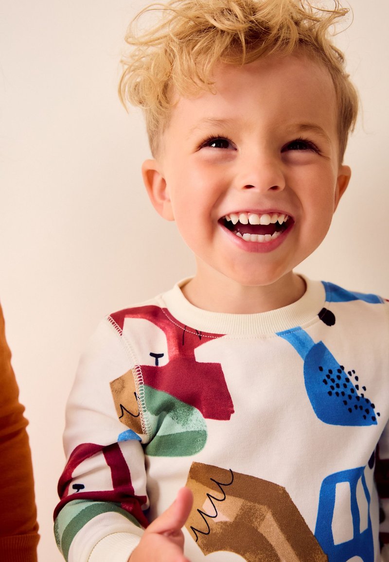 Junges Kind mit lockigem blondem Haar, das lächelt und ein farbenfrohes, gemustertes Langarmshirt vor einem neutralen Hintergrund trägt.