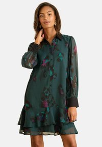 Rochie din chiffon verde cu modele florale negre și purpurii, mâneci lungi, detalii cu nasturi la manșete și tiv cu volane stratificate.