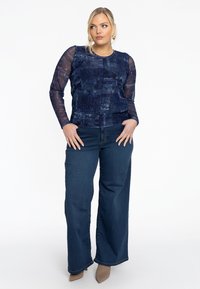 Langärmliges navy-blaues Oberteil mit Denim-Muster und durchsichtigen Ärmeln, kombiniert mit weit geschnittenen, dunklen Denim-Jeans. Die Schuhe sind hellfarbige Absatzschuhe.