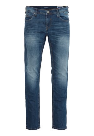 Chemise en denim bleu clair sur un t-shirt blanc avec logo noir, associée à un jean bleu et des baskets blanches ; tenue décontractée avec une coupe ample.