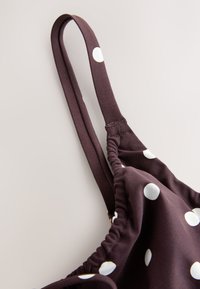 Sangle en tissu marron à pois blancs attachée à un vêtement froncé, sur un fond uni clair.