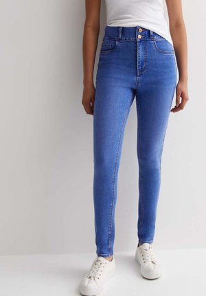 Jean skinny en denim bleu medium. Taille haute avec des accents de boutons en cuivre. Jambes ajustées avec un minimum de déchirures. Portés avec des baskets blanches.