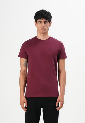 Mann mit dunklem Haar, der ein einfarbiges burgunderfarbenes T-Shirt mit kurzen Ärmeln und schwarze Hose trägt, steht vor einem weißen Hintergrund.
