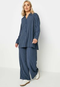 Completo blu navy a due pezzi con strisce bianche verticali, composto da una camicia con colletto e bottoni e pantaloni a gamba larga, abbinato a scarpe bianche.