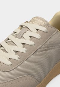 Μπεζ αθλητικό παπούτσι με υφή υφάσματος και σουέτ λεπτομέρειες, λευκά κορδόνια περασμένα από έξι οπές, και σόλα σε ανοιχτό καφέ χρώμα.