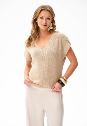 Kobieta ubrana w beżowy teksturowany top z dekoltem w serek i krótkim rękawem, jasne szerokie spodnie, duże kwiatowe kolczyki oraz masywne bransoletki na lewym nadgarstku.