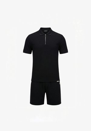 Man staat en draagt een zwarte polo met korte mouwen en rits, zwarte korte broek en zwarte sandalen tegen een effen witte achtergrond.