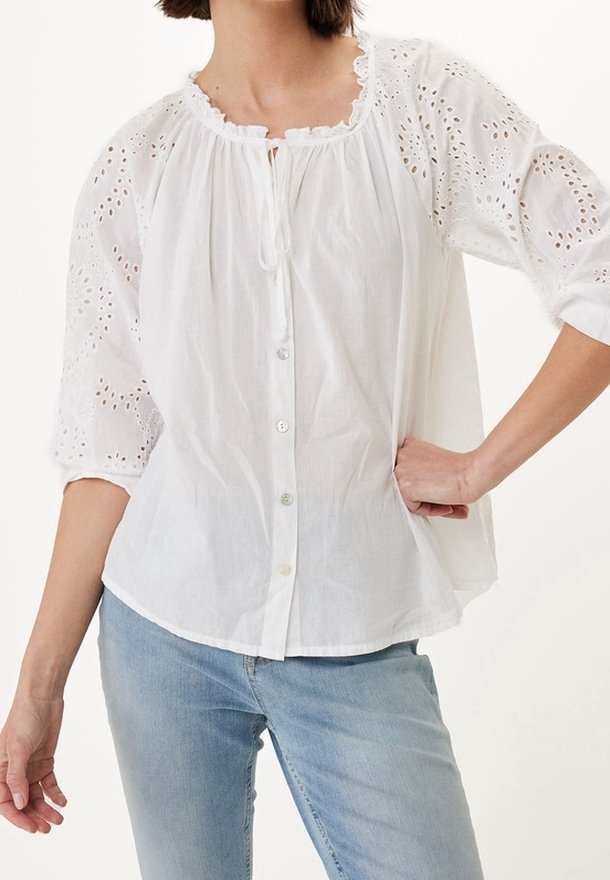 Femme portant un chemisier blanc boutonné avec des manches en dentelle ajourée et un jean bleu clair, posant avec une main sur la hanche et l'autre détendue.