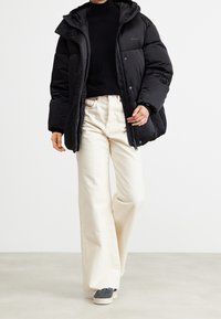 Svart pufferjacka med dragkedja fram och huva, i kombination med vita vida byxor och grå sneakers. Slät textur, minimalistisk design.