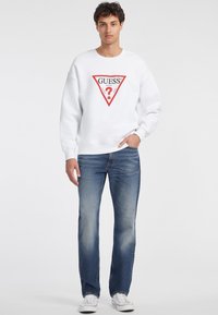 Balta sporta jaka ar sarkanu trīsstūra logo un tekstu; apvienota ar zilām džinsu biksēm un baltām sporta apaviem, stāvot uz vienkārša fona.