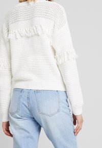 Weißer strukturierten Pullover mit offener Strickmuster, fransigen Akzenten an den Ärmeln und gerippten Bündchen, kombiniert mit hellblauen hochgradigen Jeans.